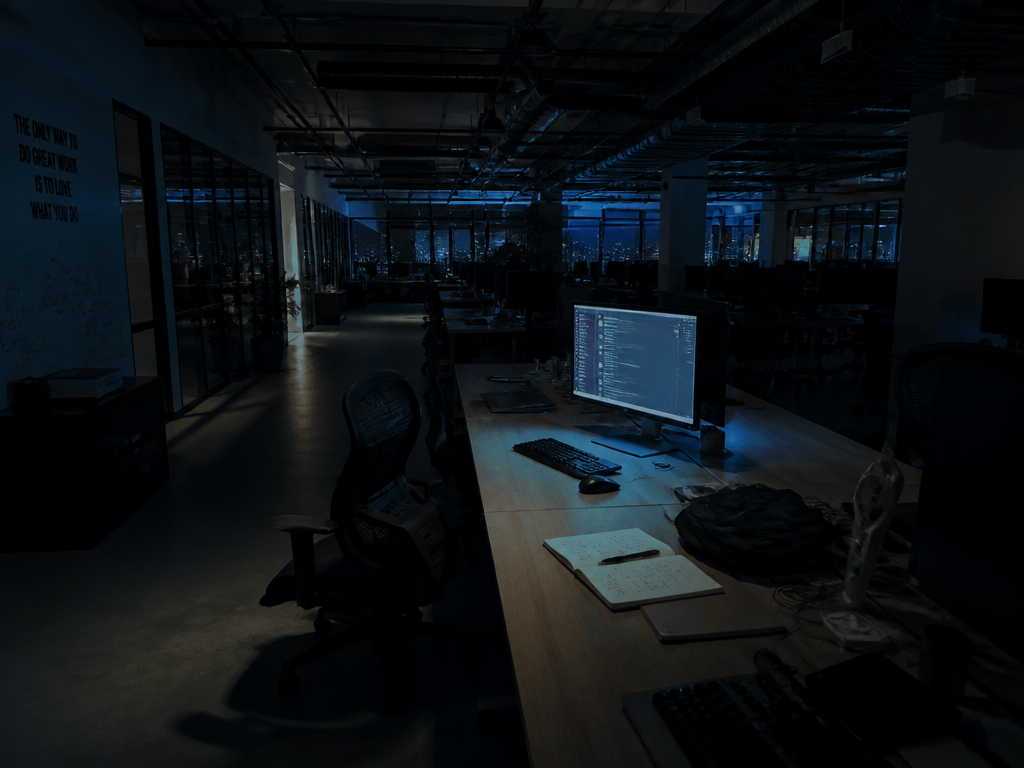A darkened open-plan startup office at night, empty ergonomic chairs pushed back from long…